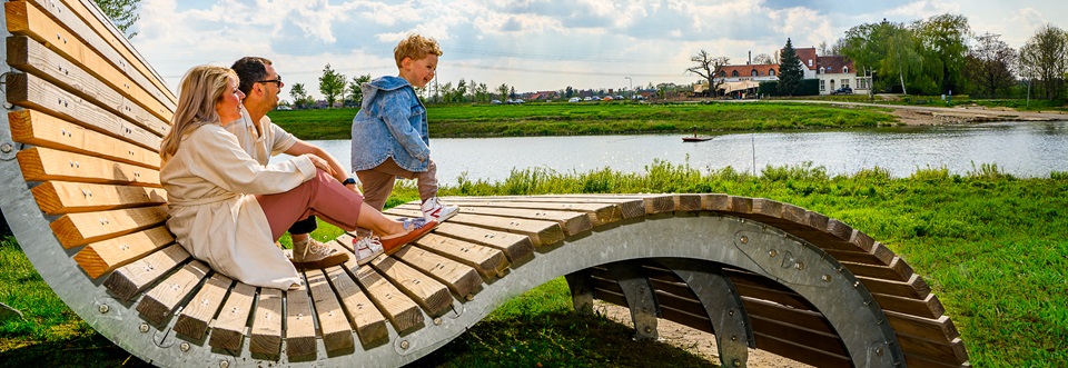 Een gezin ontspant op het golvende houten kunstwerk in Steyl langs de Maasfietsroute, met uitzicht op de Maas en het kloosterdorp aan de overkant.