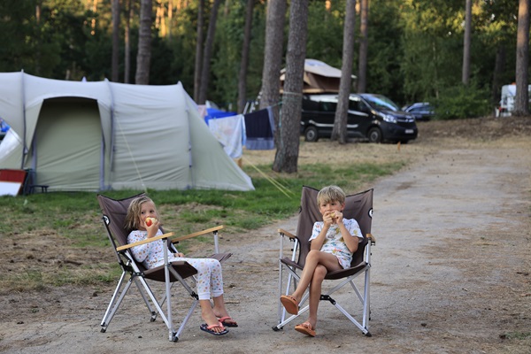 Two children sitting in camping chairs eating an apple at a campsite near Huttopia in the municipality of Roerdalen, with tents and trees in the background.